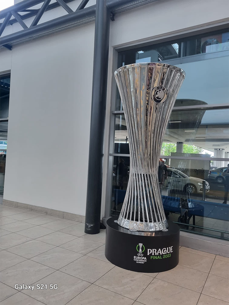 Puchar UEFA Europa Conference League stojący na czarnym stojaku z napisem PRAGUE FINAL 2023 na tle szarej ściany i okna z odbiciem samochodów.