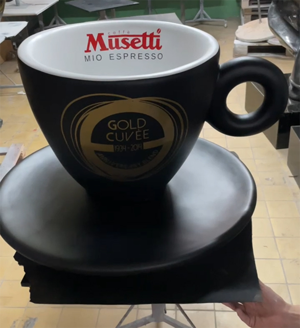 Duża czarna filiżanka kawy z napisem Musetti Mio Espresso na białym wnętrzu oraz złotym napisem Gold Cuvée 1994-2014 na zewnątrz, ustawiona na czarnym spodku, na tle pomieszczenia z jasną podłogą i stołami.