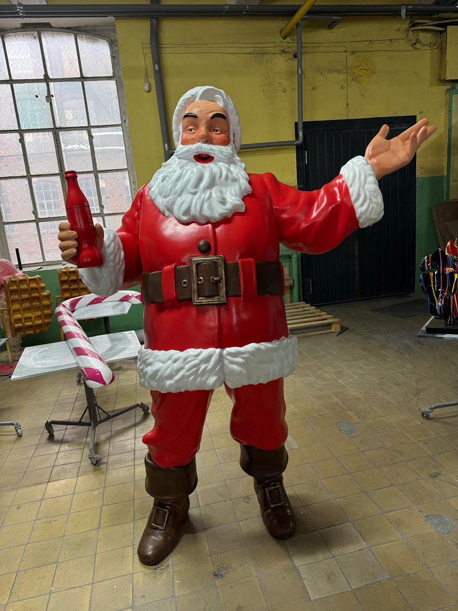 Figurka Świętego Mikołaja w czerwonym stroju i brązowych butach stojąca na podłodze w pomieszczeniu z żółto-zielonymi ścianami i dużym oknem, trzymająca czerwoną butelkę w jednej ręce i gestykulująca drugą.