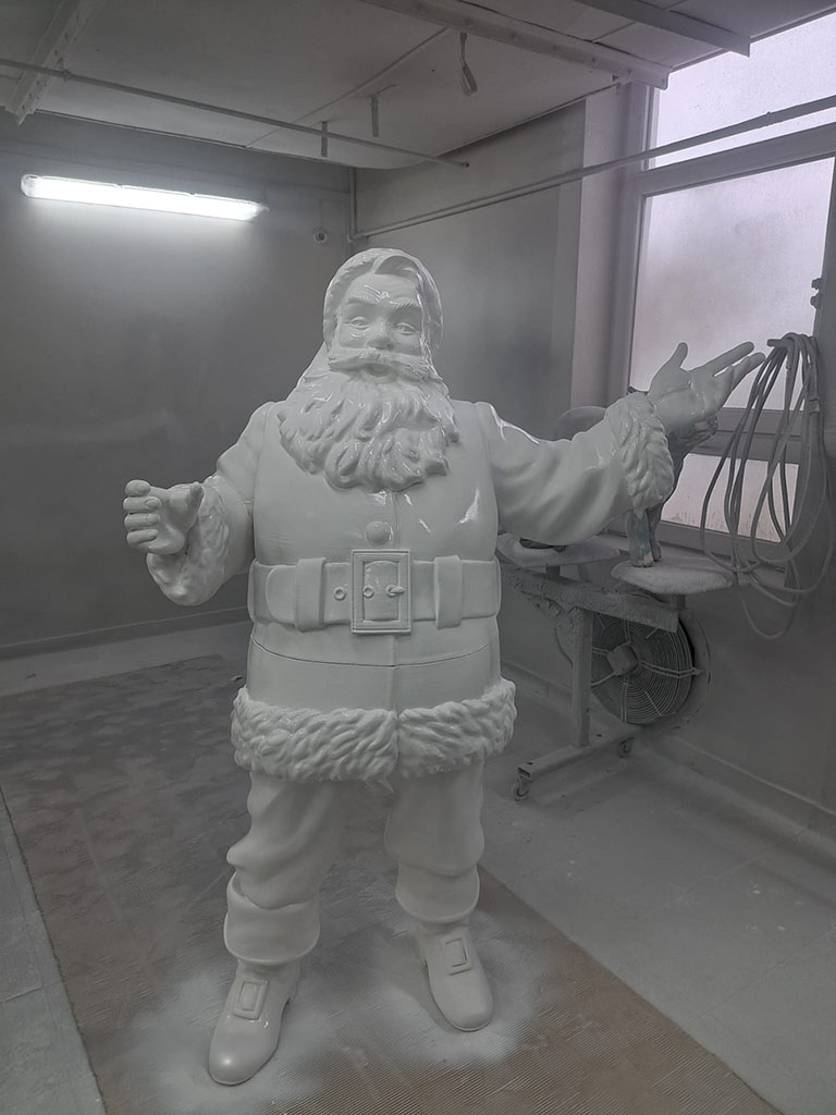 Biały, niepomalowany posąg Świętego Mikołaja w pracowni malarskiej, z uniesionymi rękoma, obok okno i wentylator.