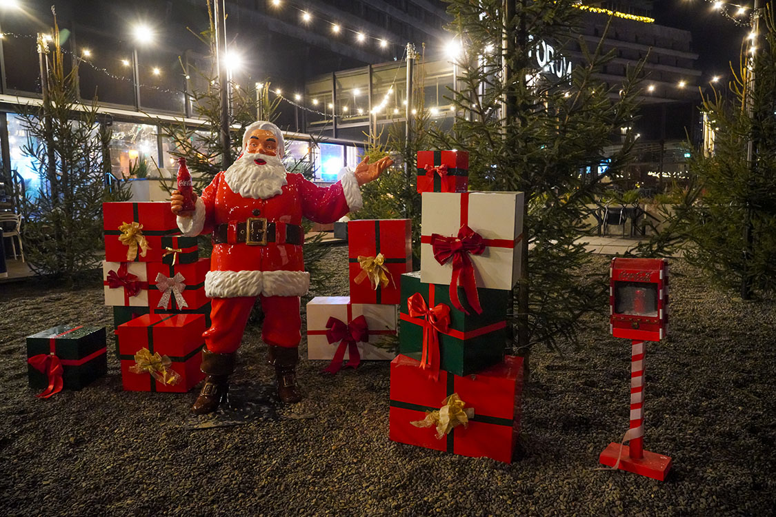 Figurka Świętego Mikołaja trzymającego butelkę Coca-Coli, otoczona licznymi prezentami w czerwonych, białych i zielonych opakowaniach z kokardami, ustawiona na żwirowym podłożu na tle drzewek iglastych i oświetlenia świątecznego nocą