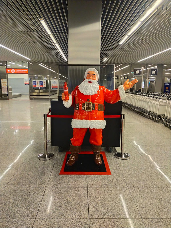 Figurka Mikołaja w czerwonym stroju z białym futrem stojąca na czarno-czerwonym podłożu, trzymająca czerwoną butelkę, w nowoczesnym hali z metalowymi sufitami i rzędem wózków po prawej stronie