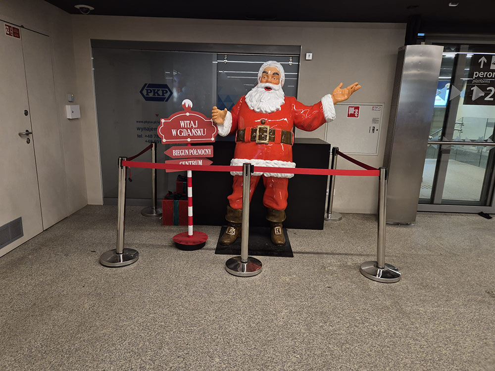 Figurka świętego Mikołaja w czerwonym stroju z białym futerkiem stojąca na dworcu w Gdańsku, obok czerwonego stojaka z napisem witającym w Gdańsku i kierunkami do Bieguna Północnego i Centrum, otoczona barierkami z czerwonym paskiem.