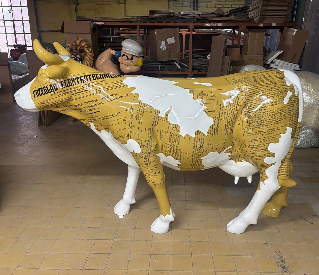 Biała i żółta rzeźba krowy pokryta tekstem i schematami technicznymi, w tle figurki postaci z kreskówek oraz stosy kartonów w magazynie.