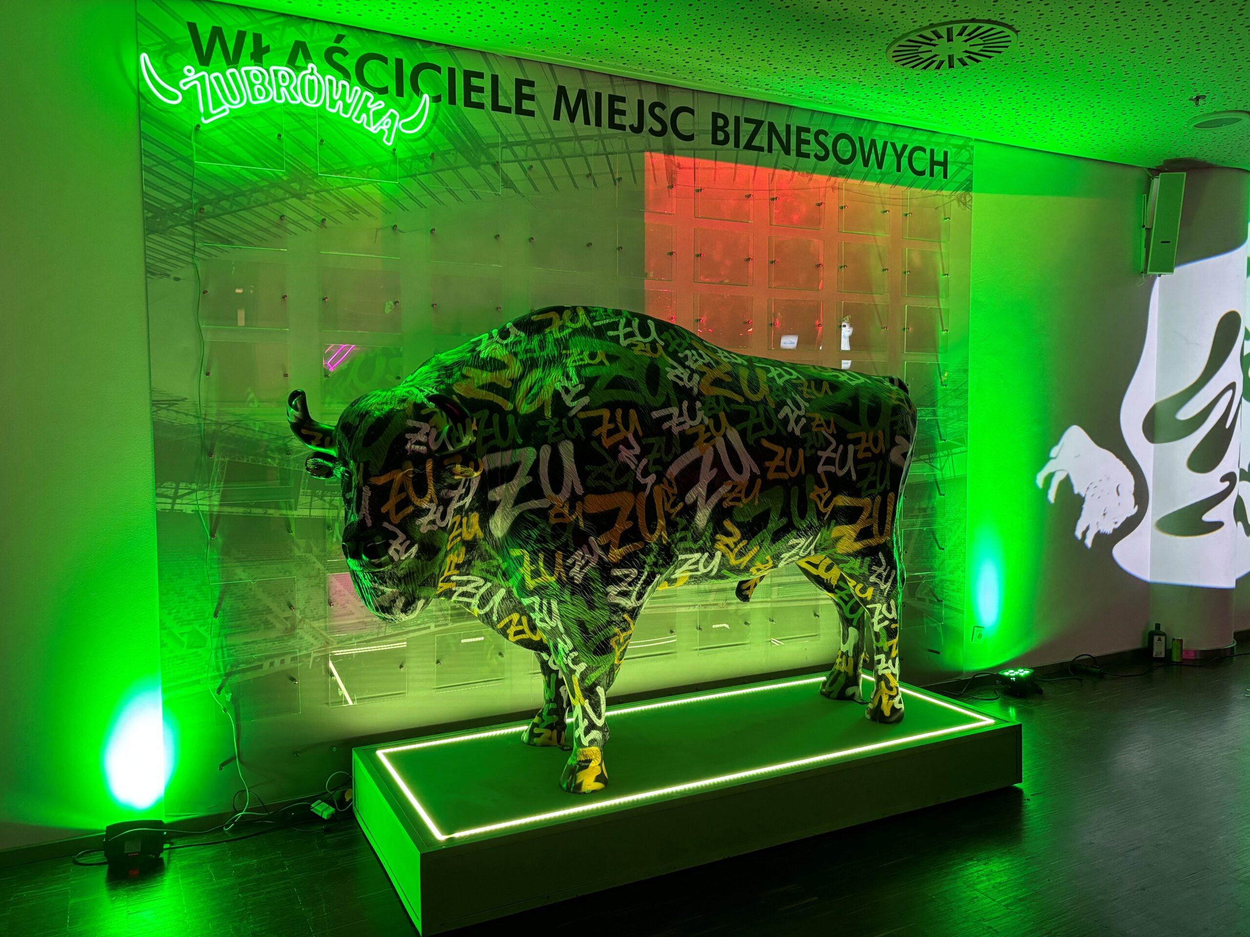 Rzeźba bizona pokryta kolorowymi napisami „ZU” na podświetlanej zielonym światłem platformie, z neonowym napisem Żubrówka i napisem na ścianie 